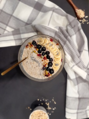 A bowl of oatmeal topped with banana slices, blackberries, nuts, and seeds is placed on a table. A white and gray checkered cloth is draped around it. Adjacent to the bowl, there is a wooden spoon with scattered oats.