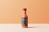 A single sauce bottle with an orange hue is centrally placed against a smooth, gradient orange background. The bottle is topped with a brown cap and features a dark label with white text. The label includes the words 'SAUCE BOTTLE' prominently.