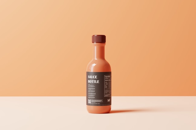 A single sauce bottle with an orange hue is centrally placed against a smooth, gradient orange background. The bottle is topped with a brown cap and features a dark label with white text. The label includes the words 'SAUCE BOTTLE' prominently.