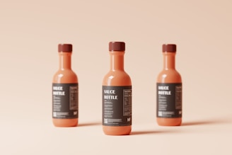Three orange-colored sauce bottles with dark labels are arranged on a light peach background. The bottles are symmetrical and evenly spaced, casting soft shadows.