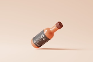 A small, glass bottle filled with a reddish-orange sauce is suspended in mid-air against a beige background. The bottle has a dark label with white text that reads 'SAUCE BOTTLE' and contains other small details. Its cap matches the dark label color.