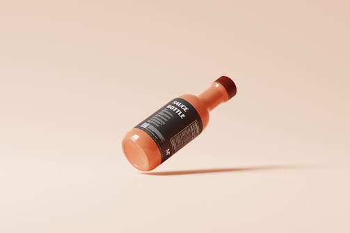 A sauce bottle with a black label is suspended mid-air against a minimalist beige background. The label contains text in white, and the bottle appears to be sealed with a matching cap. The overall design is clean and modern.