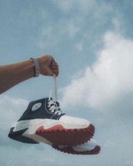 An arm wearing a watch is holding a pair of sneakers with white laces against a backdrop of a cloudy blue sky. The sneakers have a black and white design with distinct red soles.