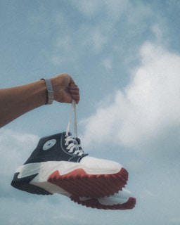An arm wearing a watch is holding a pair of sneakers with white laces against a backdrop of a cloudy blue sky. The sneakers have a black and white design with distinct red soles.