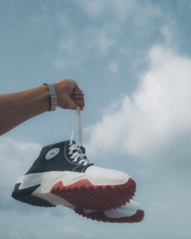 An arm wearing a watch is holding a pair of sneakers with white laces against a backdrop of a cloudy blue sky. The sneakers have a black and white design with distinct red soles.
