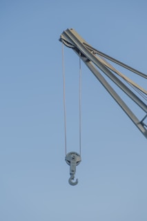A close-up view of a crane with a metal hook hanging from it against a clear blue sky. The crane is industrial in design, made of sturdy metal with ropes or cables for lifting heavy objects.