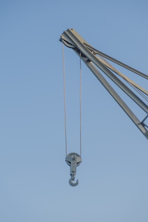 A close-up view of a crane with a metal hook hanging from it against a clear blue sky. The crane is industrial in design, made of sturdy metal with ropes or cables for lifting heavy objects.