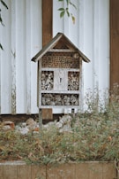 A wooden insect hotel with a rustic design is positioned against a white plank wall. The structure includes various natural materials like pinecones, hollow logs, and twigs, providing shelter for insects. It is surrounded by overgrown grass and plants, adding to the natural setting.