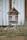 A wooden insect hotel with a rustic design is positioned against a white plank wall. The structure includes various natural materials like pinecones, hollow logs, and twigs, providing shelter for insects. It is surrounded by overgrown grass and plants, adding to the natural setting.