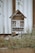 A wooden insect hotel with a rustic design is positioned against a white plank wall. The structure includes various natural materials like pinecones, hollow logs, and twigs, providing shelter for insects. It is surrounded by overgrown grass and plants, adding to the natural setting.