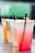 A row of colorful beverages in transparent plastic cups with black straws, placed on a metal table with a perforated pattern. The drinks range from orange and greenish hues to light yellow and pink tones. The background is blurred, highlighting the vibrant colors of the drinks.
