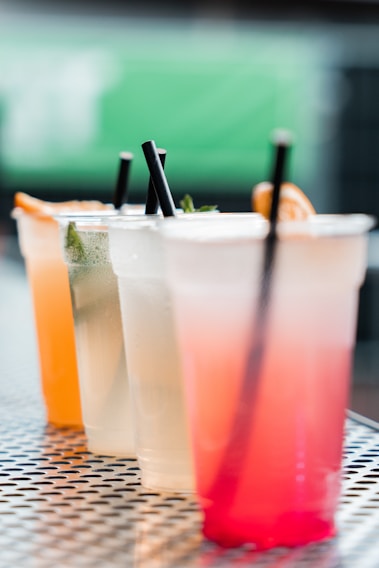 A row of colorful beverages in transparent plastic cups with black straws, placed on a metal table with a perforated pattern. The drinks range from orange and greenish hues to light yellow and pink tones. The background is blurred, highlighting the vibrant colors of the drinks.