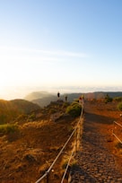 A scenic landscape with a cobblestone path leading to a viewpoint. Several people are standing on the platform, overlooking a vast expanse of rolling hills and valleys. Sunlight casts a warm glow over the landscape, highlighting the greenery and rocky terrain.
