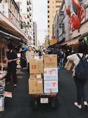 A bustling urban street market with various stalls and people walking. Cardboard boxes are stacked on a cart in the center, and signs with Japanese text are visible on the buildings. A large red crab decoration is mounted on a building facade.