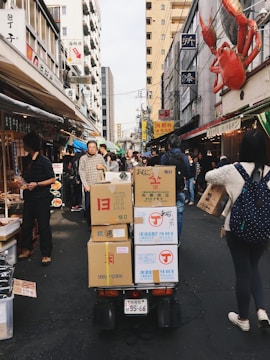 A bustling urban street market with various stalls and people walking. Cardboard boxes are stacked on a cart in the center, and signs with Japanese text are visible on the buildings. A large red crab decoration is mounted on a building facade.