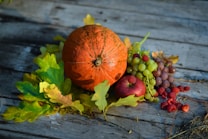 A collection of autumn harvest items is arranged on a wooden surface. A large orange pumpkin sits next to vibrant green and yellow leaves. Nearby, there are bunches of grapes in both green and purple hues, alongside a bright red pomegranate and scattered red raspberries, creating a visually appealing display.