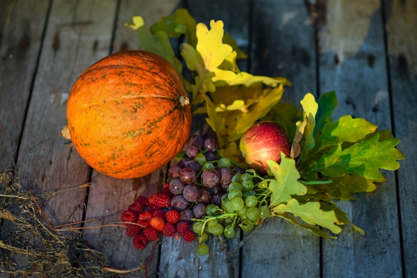 A collection of autumn fruits and foliage placed on a rustic wooden surface. The arrangement includes an orange pumpkin, a red apple, clusters of purple and green grapes, red raspberries, and oak leaves with varying shades of green and yellow.