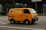 A bright yellow delivery van is driving down a city street. The van has the text 'FRESH DELI' and a phone number on its side. The background features a modern glass building and some traffic signs. The road is empty except for the van.