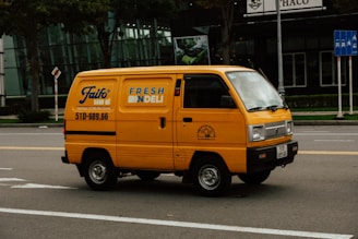 A bright yellow delivery van is driving down a city street. The van has the text 'FRESH DELI' and a phone number on its side. The background features a modern glass building and some traffic signs. The road is empty except for the van.
