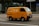 A bright yellow delivery van is driving down a city street. The van has the text 'FRESH DELI' and a phone number on its side. The background features a modern glass building and some traffic signs. The road is empty except for the van.