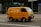 A bright yellow delivery van is driving down a city street. The van has the text 'FRESH DELI' and a phone number on its side. The background features a modern glass building and some traffic signs. The road is empty except for the van.