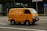 A bright yellow delivery van is driving down a city street. The van has the text 'FRESH DELI' and a phone number on its side. The background features a modern glass building and some traffic signs. The road is empty except for the van.