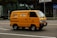 A bright yellow delivery van is driving down a city street. The van has the text 'FRESH DELI' and a phone number on its side. The background features a modern glass building and some traffic signs. The road is empty except for the van.