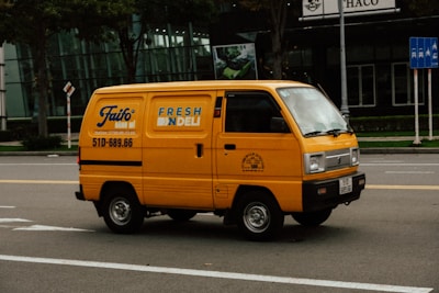 A bright yellow delivery van is driving down a city street. The van has the text 'FRESH DELI' and a phone number on its side. The background features a modern glass building and some traffic signs. The road is empty except for the van.