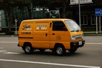 A bright yellow delivery van is driving down a city street. The van has the text 'FRESH DELI' and a phone number on its side. The background features a modern glass building and some traffic signs. The road is empty except for the van.