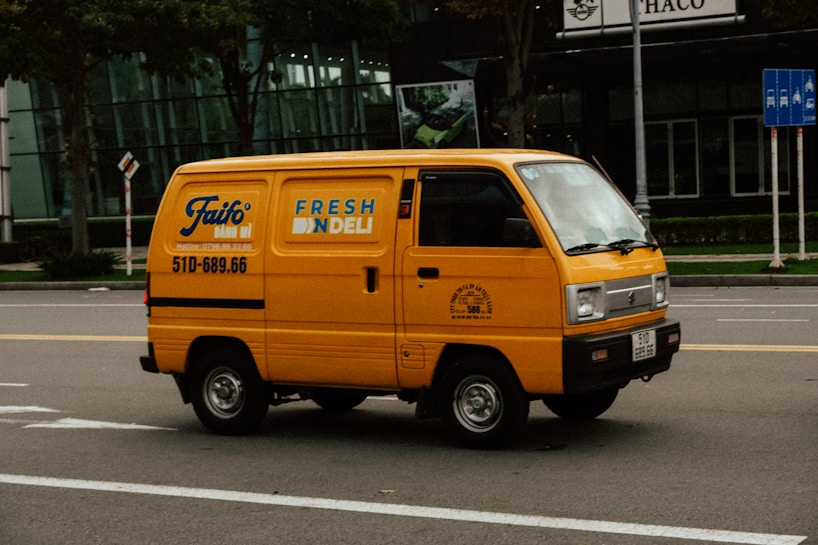 A bright yellow delivery van is driving down a city street. The van has the text 'FRESH DELI' and a phone number on its side. The background features a modern glass building and some traffic signs. The road is empty except for the van.