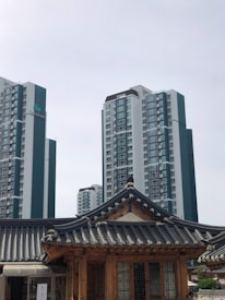 A modern cityscape combines traditional and contemporary architecture. In the foreground, a wooden structure features a traditional tiled roof, exemplifying classical design elements. In the background, high-rise apartment buildings rise into the overcast sky, showcasing modern urban development.
