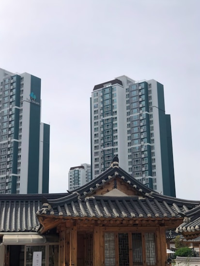 A modern cityscape combines traditional and contemporary architecture. In the foreground, a wooden structure features a traditional tiled roof, exemplifying classical design elements. In the background, high-rise apartment buildings rise into the overcast sky, showcasing modern urban development.