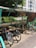 A covered bicycle parking area with several bikes parked under a metal and glass shelter. The area is shaded by trees on one side, and a multi-story building is visible in the background. Sunlight filters through the foliage, casting dappled shadows on the ground.