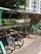 A covered bicycle parking area with several bikes parked under a metal and glass shelter. The area is shaded by trees on one side, and a multi-story building is visible in the background. Sunlight filters through the foliage, casting dappled shadows on the ground.