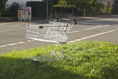 A metal shopping cart is resting on a patch of grass beside a road. The surrounding environment includes a street with road markings and some greenery, including bushes and lawns. In the background, a small electric scooter is parked near a utility box covered with graffiti.