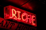 A vibrant red neon sign displaying the word 'RICHE', mounted against a dark background. The letters are illuminated in a bold and striking font, casting a warm glow.
