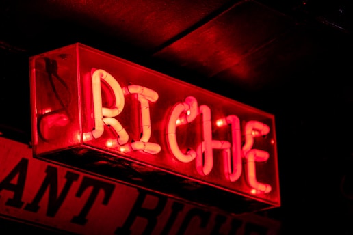 A vibrant red neon sign displaying the word 'RICHE', mounted against a dark background. The letters are illuminated in a bold and striking font, casting a warm glow.