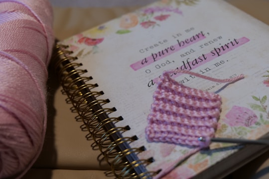 A spiral-bound notebook is lying open with a decorative floral design and a biblical quote printed on one page. Next to it, there is a partial piece of pink crochet with a metal crochet hook and a skein of pink yarn.