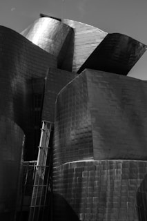 A complex structure composed of interlocking metallic surfaces that reflect light in varying tones of gray. The angular and curved forms create an interplay of shadows and highlights on the reflective metal, lending an abstract and modern architectural aesthetic.