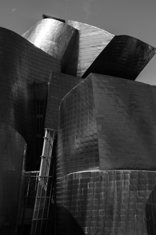 A complex structure composed of interlocking metallic surfaces that reflect light in varying tones of gray. The angular and curved forms create an interplay of shadows and highlights on the reflective metal, lending an abstract and modern architectural aesthetic.