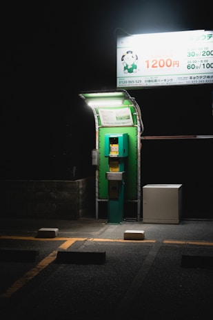 A dimly lit parking payment machine is situated under a bright overhead sign on a dark night. The machine is green and stands next to a white utility box. The pavement is marked with yellow lines and parking blocks, with an overall atmosphere of solitude and quiet.