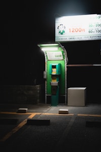 A dimly lit parking payment machine is situated under a bright overhead sign on a dark night. The machine is green and stands next to a white utility box. The pavement is marked with yellow lines and parking blocks, with an overall atmosphere of solitude and quiet.