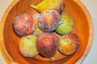 A wooden bowl holds several figs with a mix of reddish-purple and greenish-yellow colors. The figs are arranged with some facing up, displaying their tops and stems, while others rest on their sides.