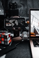A cluttered desktop setup features a smartphone displaying the time and location, mounted on a bear-shaped stand. Beside it, a Rubik's cube adds a pop of color to the scene. A small figurine resembling a cartoon platypus sits nearby, alongside part of an open laptop showing architectural imagery. The background consists of a quilted gray surface with various electronic components.