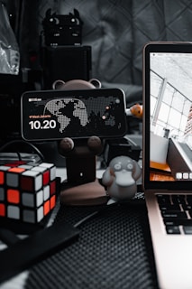 A cluttered desktop setup features a smartphone displaying the time and location, mounted on a bear-shaped stand. Beside it, a Rubik's cube adds a pop of color to the scene. A small figurine resembling a cartoon platypus sits nearby, alongside part of an open laptop showing architectural imagery. The background consists of a quilted gray surface with various electronic components.