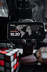 A smartphone displays a clock with the time 10:20, placed on a holder with animal paw designs. There's a map in the background and the location is set to West Jakarta. On the left, a Rubik's cube is visible, and on the right, a figure resembling a cartoon duck-like character is present. The background is filled with electronic equipment.