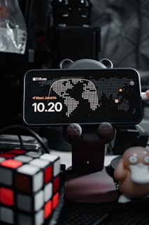A smartphone displays a clock with the time 10:20, placed on a holder with animal paw designs. There's a map in the background and the location is set to West Jakarta. On the left, a Rubik's cube is visible, and on the right, a figure resembling a cartoon duck-like character is present. The background is filled with electronic equipment.