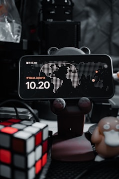 A smartphone displays a clock with the time 10:20, placed on a holder with animal paw designs. There's a map in the background and the location is set to West Jakarta. On the left, a Rubik's cube is visible, and on the right, a figure resembling a cartoon duck-like character is present. The background is filled with electronic equipment.