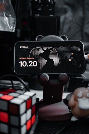 A smartphone displays a clock with the time 10:20, placed on a holder with animal paw designs. There's a map in the background and the location is set to West Jakarta. On the left, a Rubik's cube is visible, and on the right, a figure resembling a cartoon duck-like character is present. The background is filled with electronic equipment.