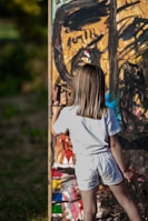 A young child with shoulder-length hair is standing and painting on a large, colorful canvas outdoors. The artwork consists of bold, abstract strokes in a variety of colors including black, orange, blue, and red. The child is wearing a white shirt and shorts, and appears to be focused on their creative task.